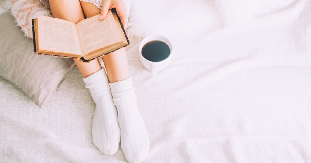 Woman sitting in bed with socks on her feet while reading a book.
