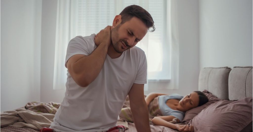 Man holding his neck as he sits on his bed